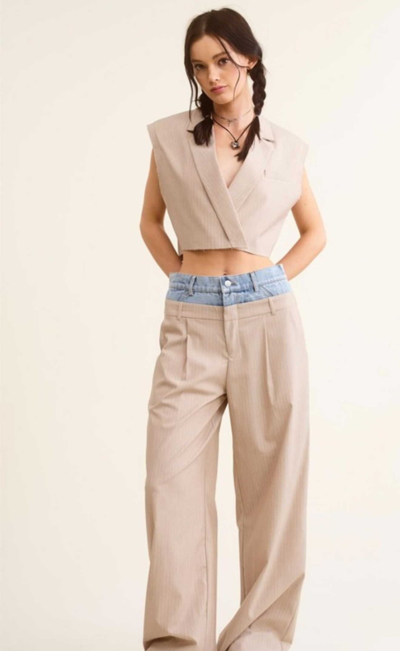 Woman wearing a beige sleeveless blazer and high-waisted pants with a blue belt on a plain background