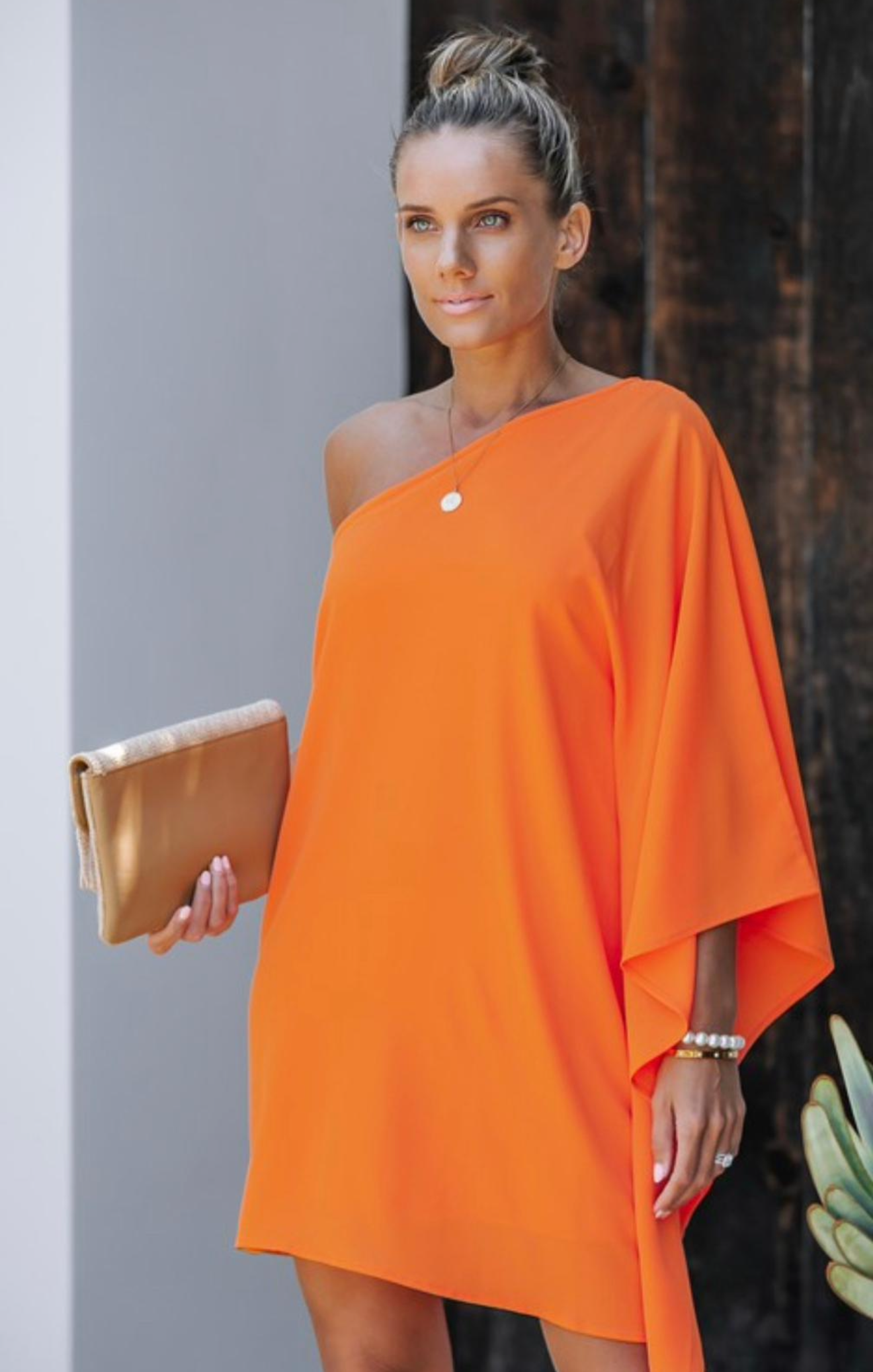 Woman wearing an orange one-shoulder dress holding a clutch against a neutral background